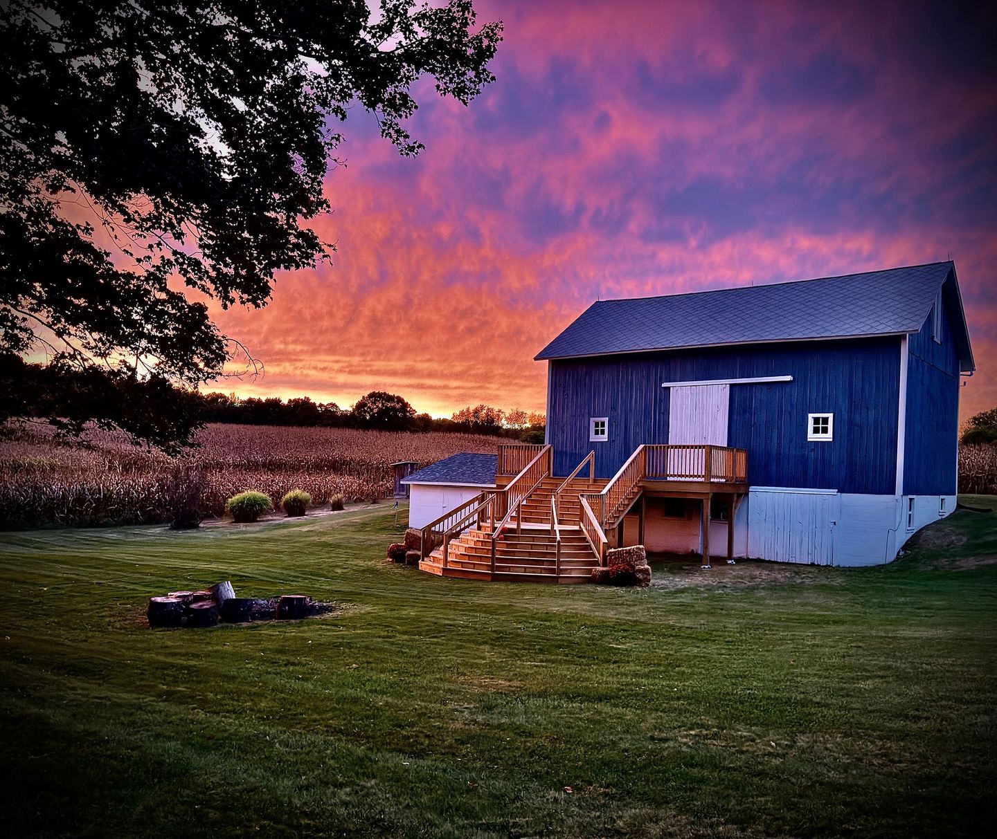 Blue barn wedding venue with wooden deck and stairs, set against a vibrant sunset sky over a green field.
