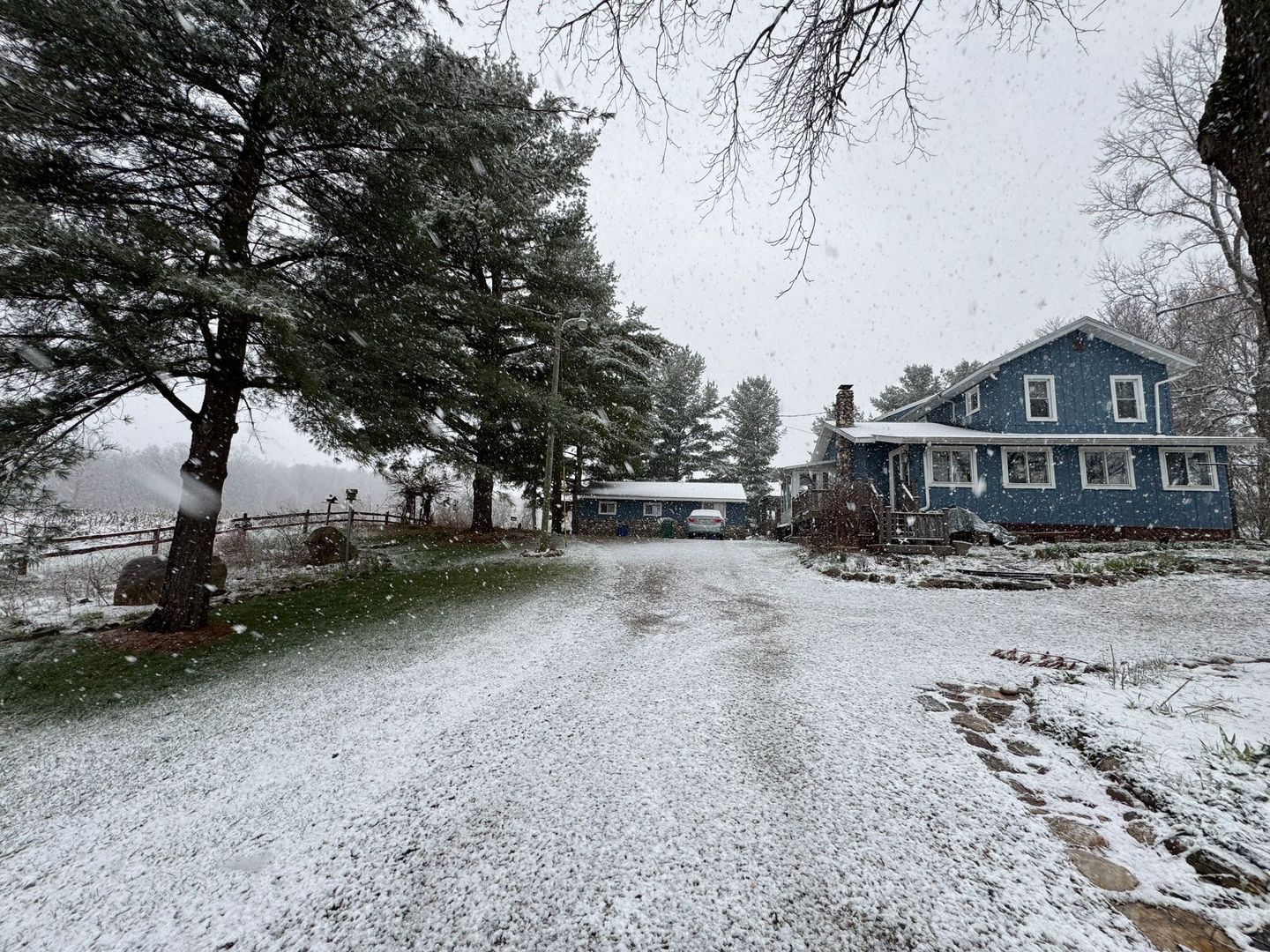 Snowy scene at Inn Between the Lakes with a blue two-story farm house, driveway, and trees. Snowflakes fall.