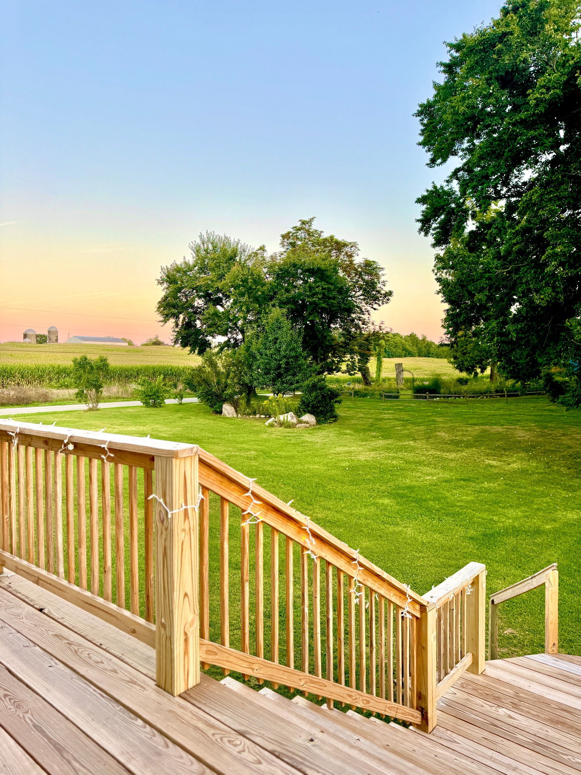 Wooden deck stairs lead to a green lawn with trees, fields, and a sunset sky.