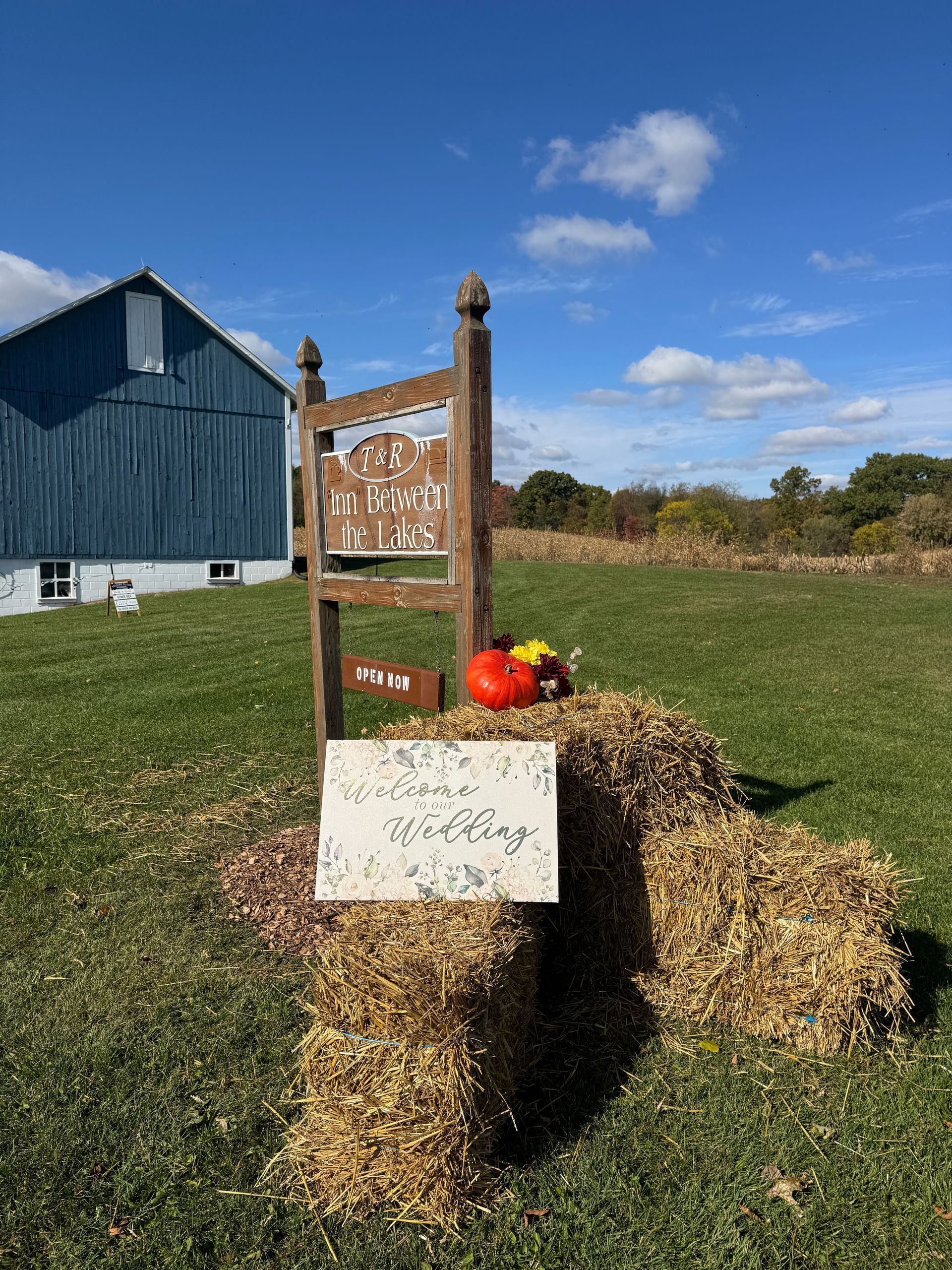 Straw bales with pumpkins and an event sign, set on hay bales in front of a blue wedding and event venue barn.