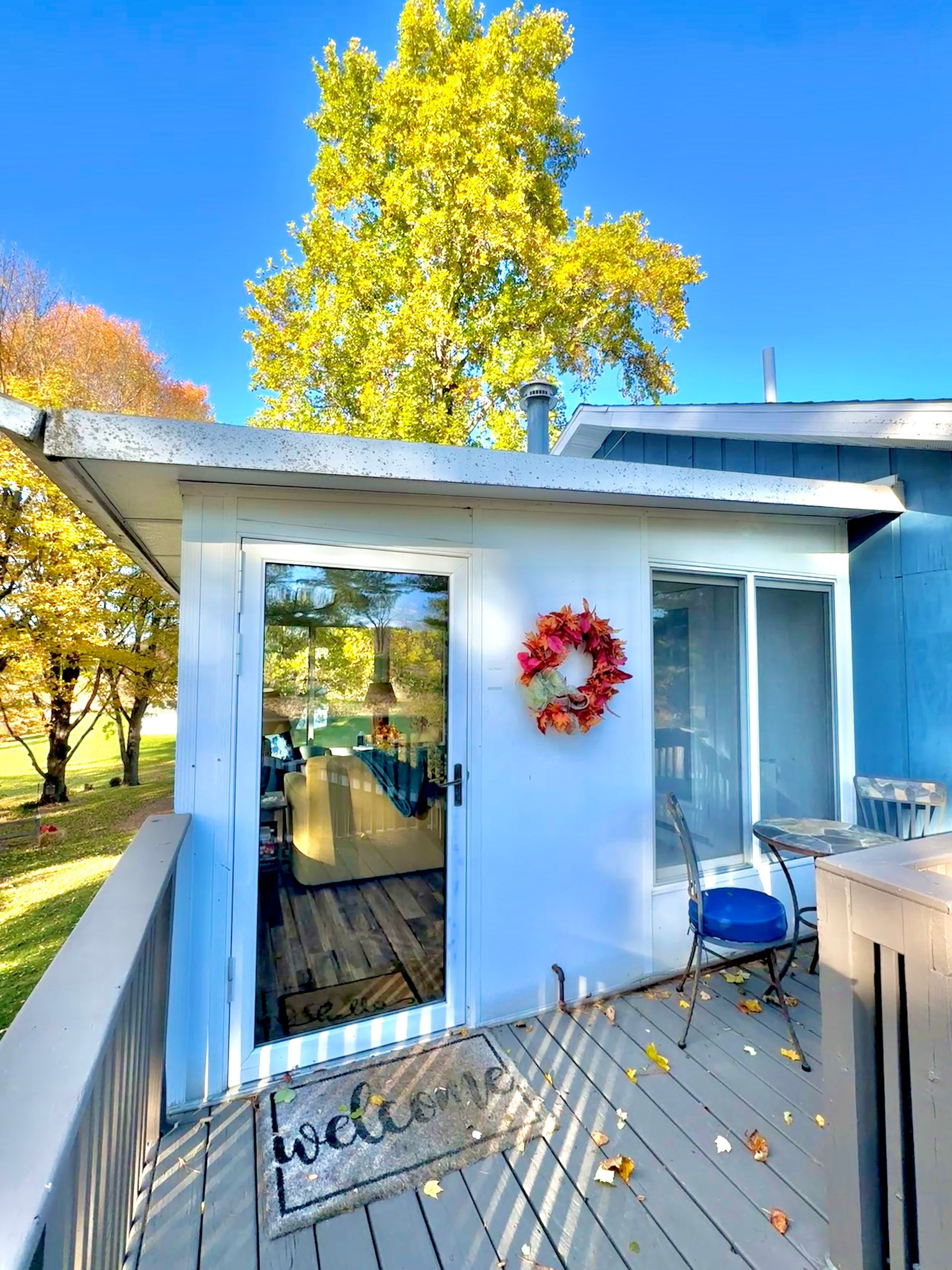 White-walled porch with a glass door, fall wreath, table, chair, and a large tree with yellow leaves under a blue sky.
