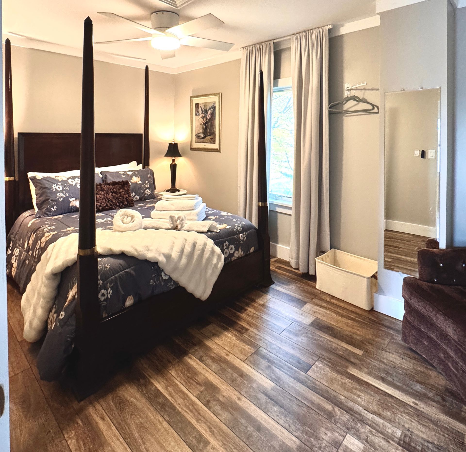 Bedroom with four-poster queen bed, wood floor, mirror, window with curtains, and a white ceiling fan.