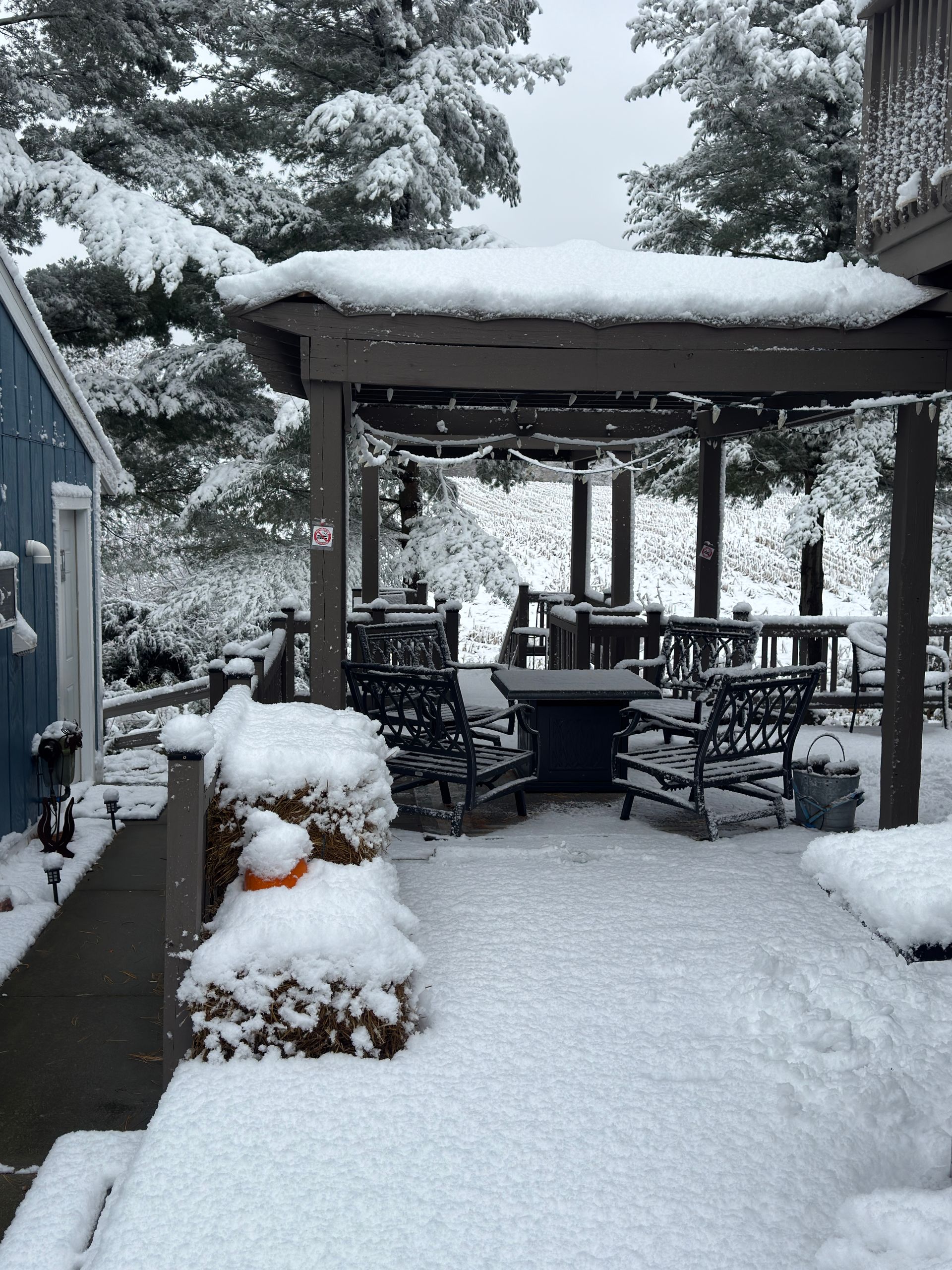 Snow-covered outdoor patio with pergola, chairs, and tables. Snow blankets the ground, trees, and structures.