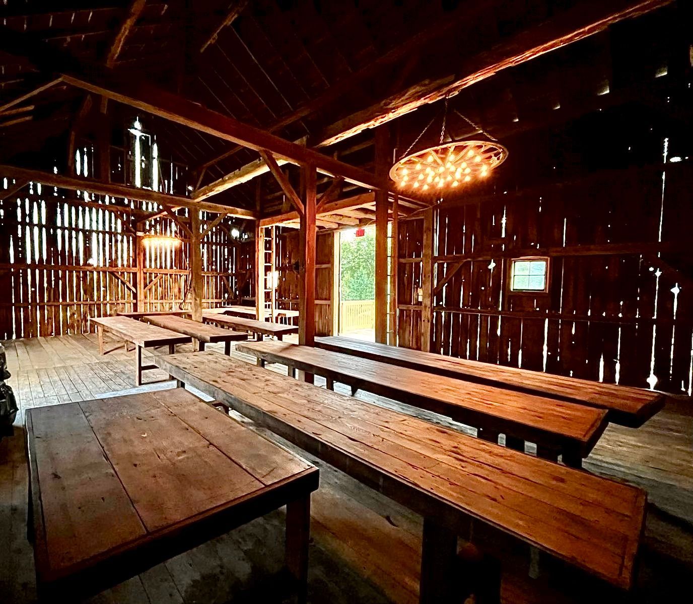 Rustic wedding and event venue barn interior with long wooden tables. Dimly lit with hanging wagon wheel lights and natural light from openings.