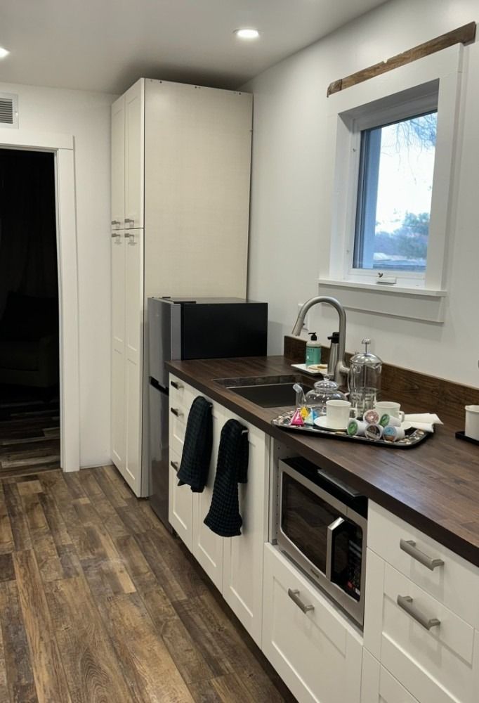 Kitchenette with white cabinets, dark countertop, sink, microwave, window, refrigerator, and doorway.