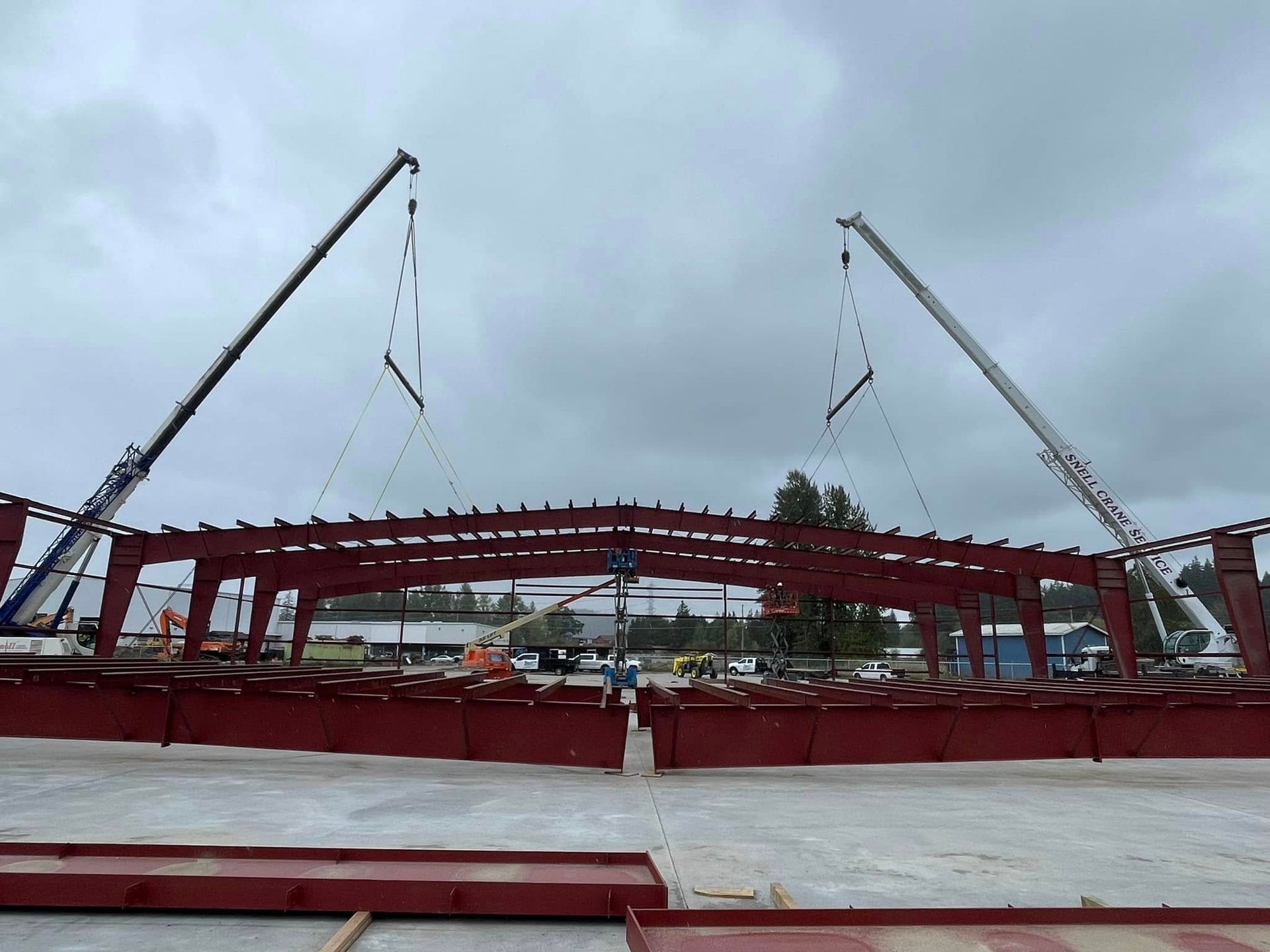 Two cranes hoisting a red steel beam structure at a construction site