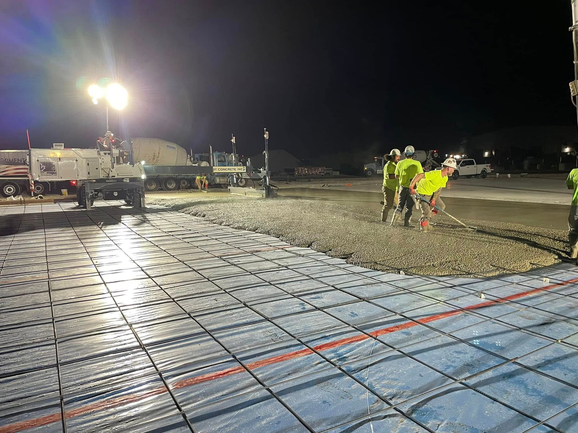 Nighttime construction site with workers in neon vests on a concrete slab under bright floodlights