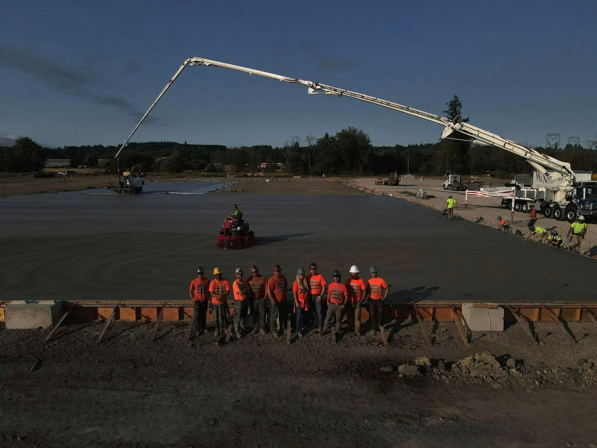 Crew in orange vests on a muddy construction site beside a long concrete pump boom.