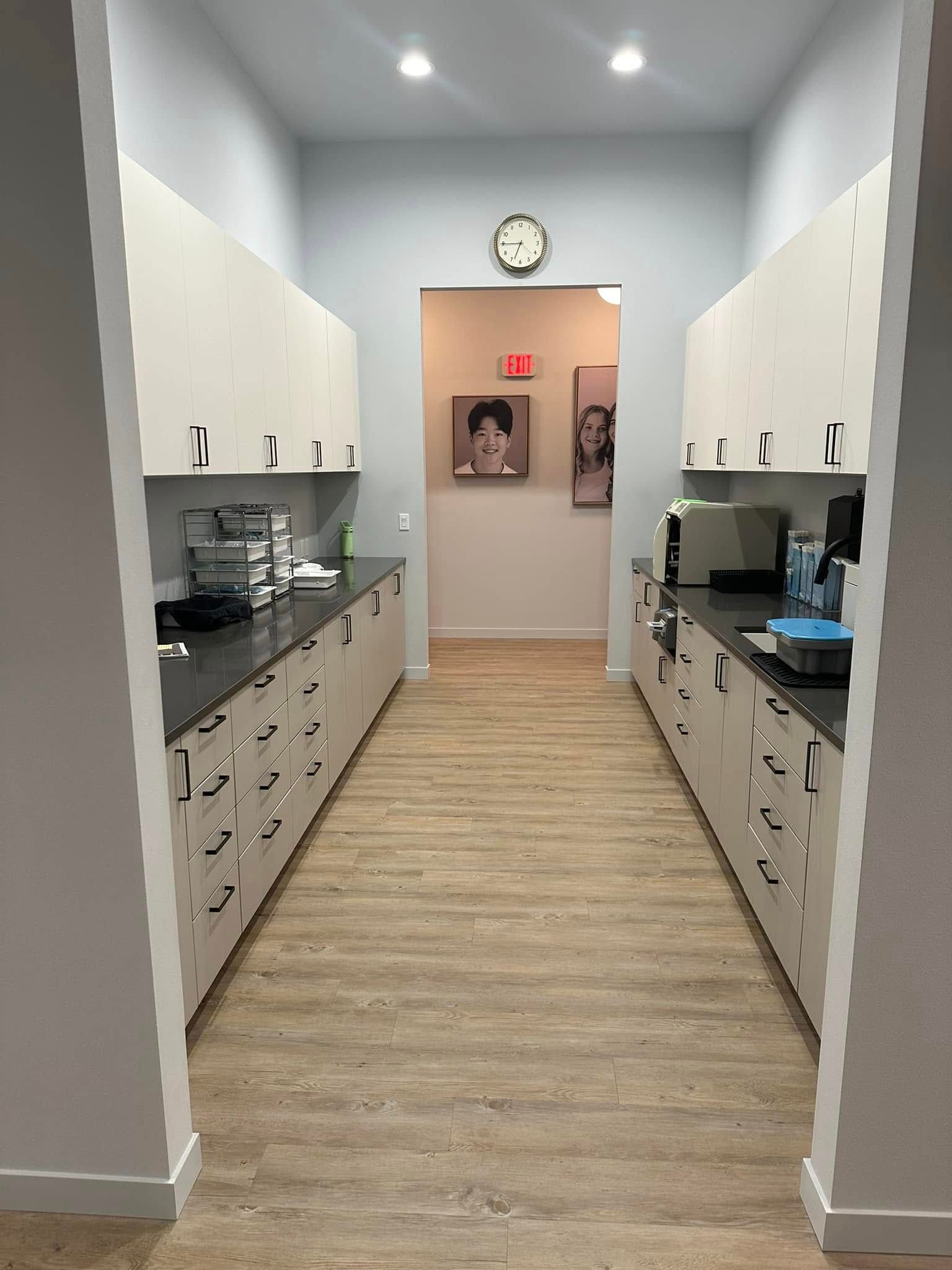 Narrow kitchen hallway with white cabinets, wood floor, and a clock above a door at the end.