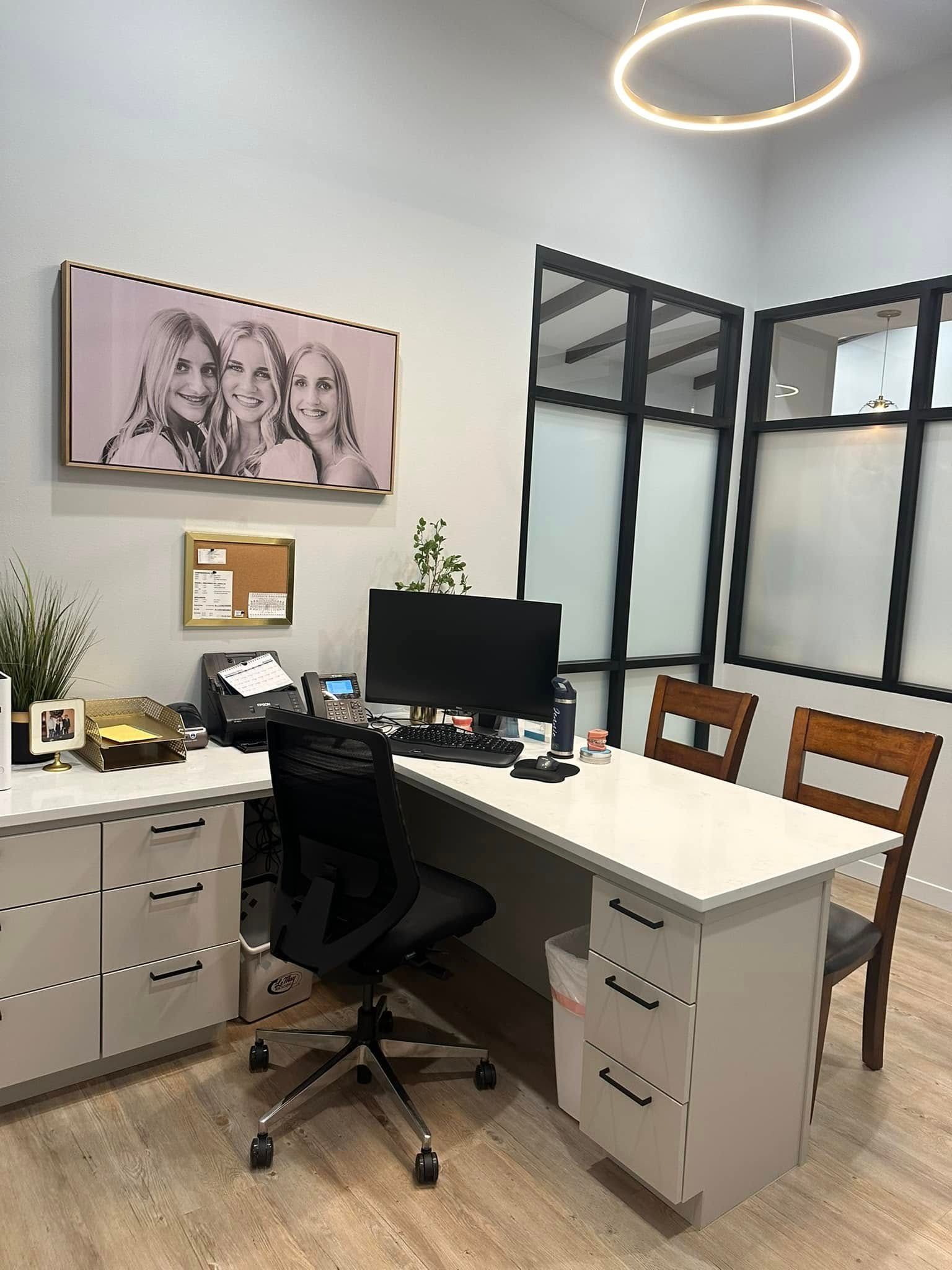 Modern office corner with white desk, computer monitor, black chair, cabinets, wall art, and glass partitions