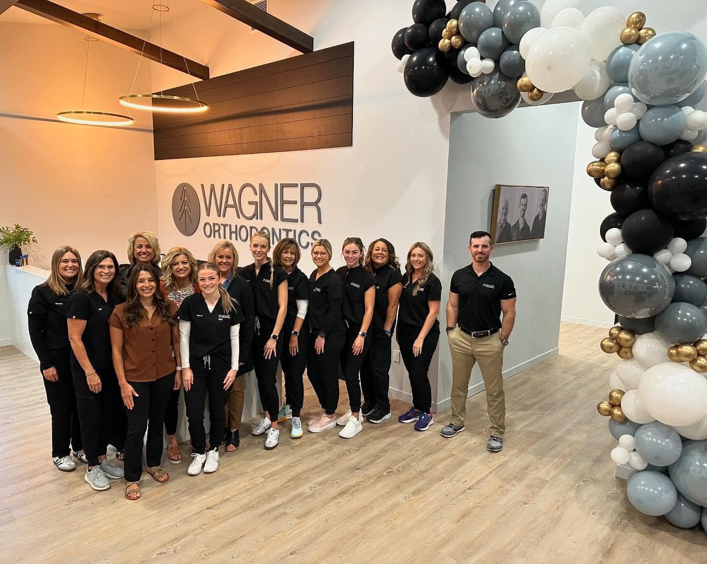 Group of staff posing at Wagner Orthodontics under black, white, and gray balloon arches in a lobby