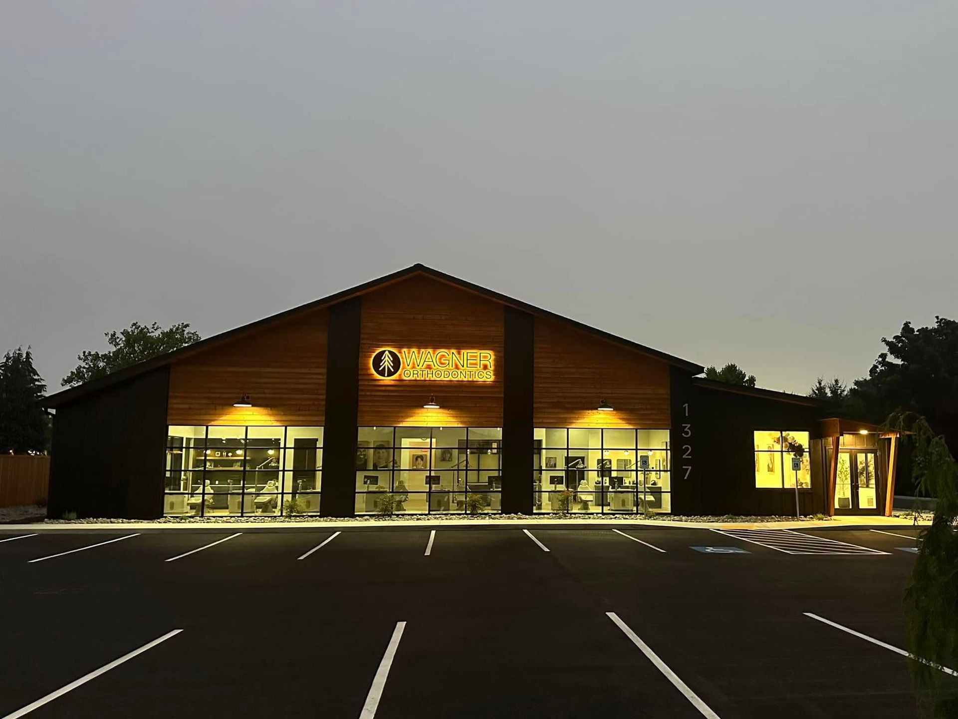 A lit storefront with a large peaked roof at dusk, seen across an empty parking lot.