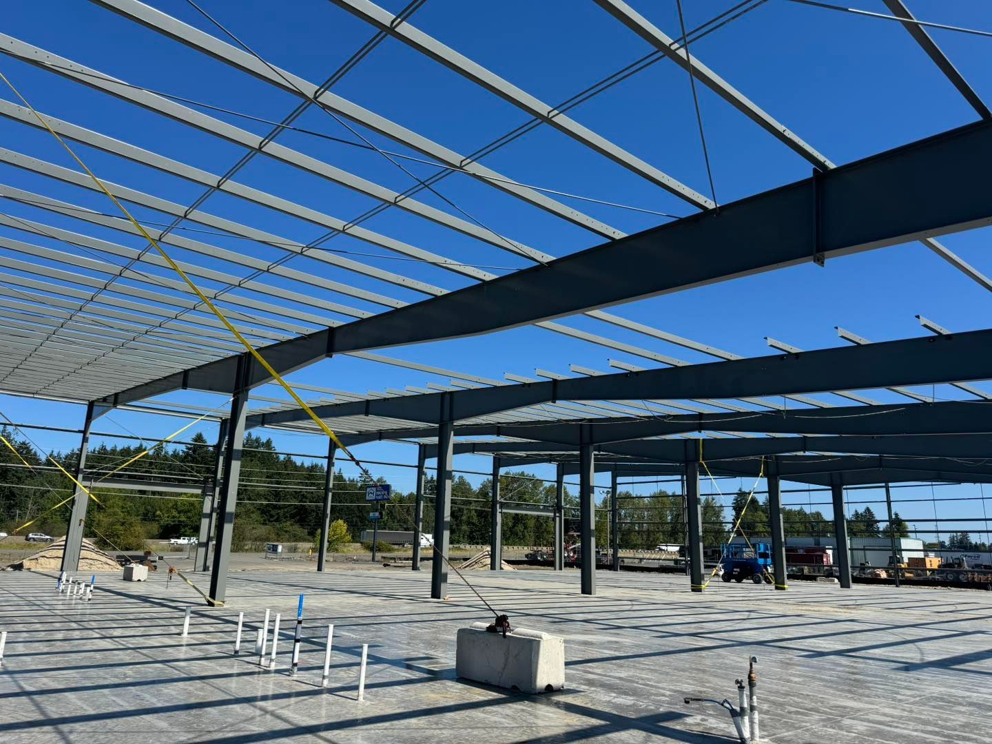 Partially built steel structure with roof beams on a concrete lot under a clear blue sky