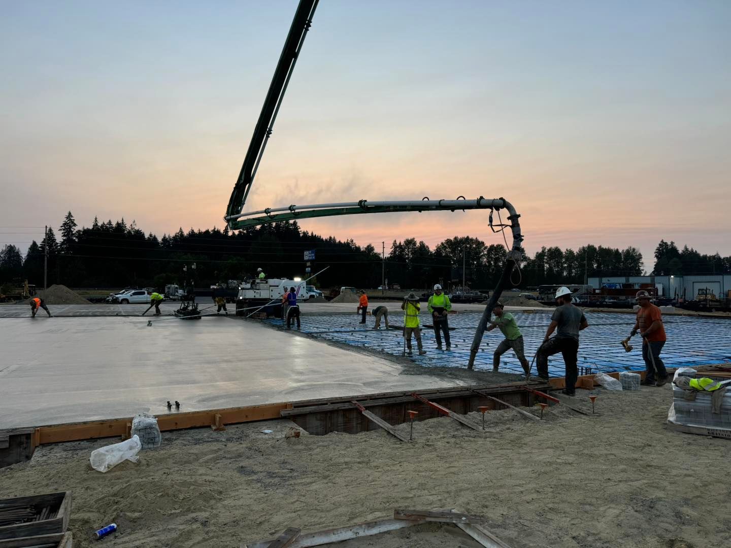 Workers pouring concrete at dusk while a long boom pump stretches over a large slab foundation.