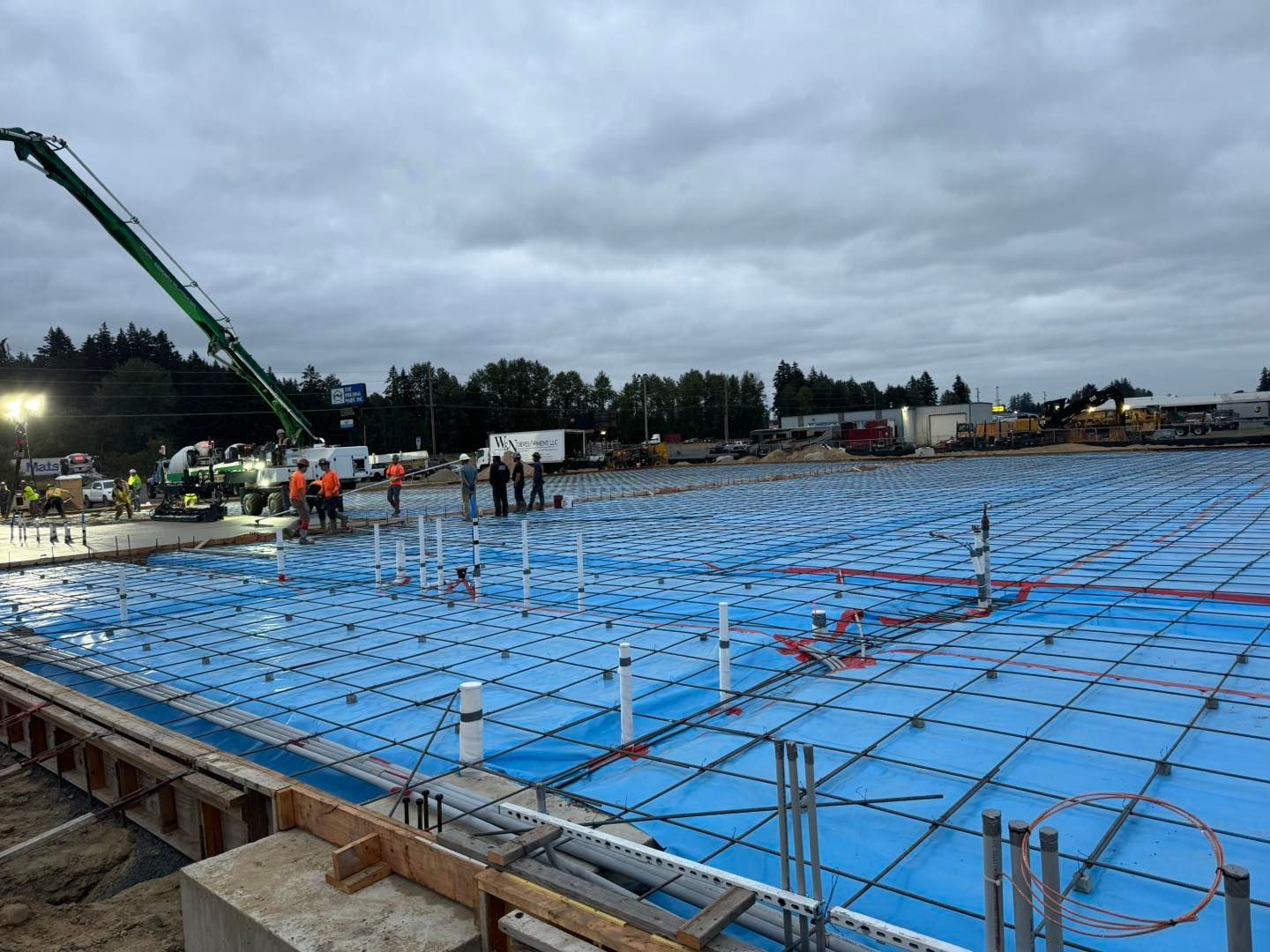 Construction site with a large blue tarp-covered foundation, workers, and a crane under cloudy skies
