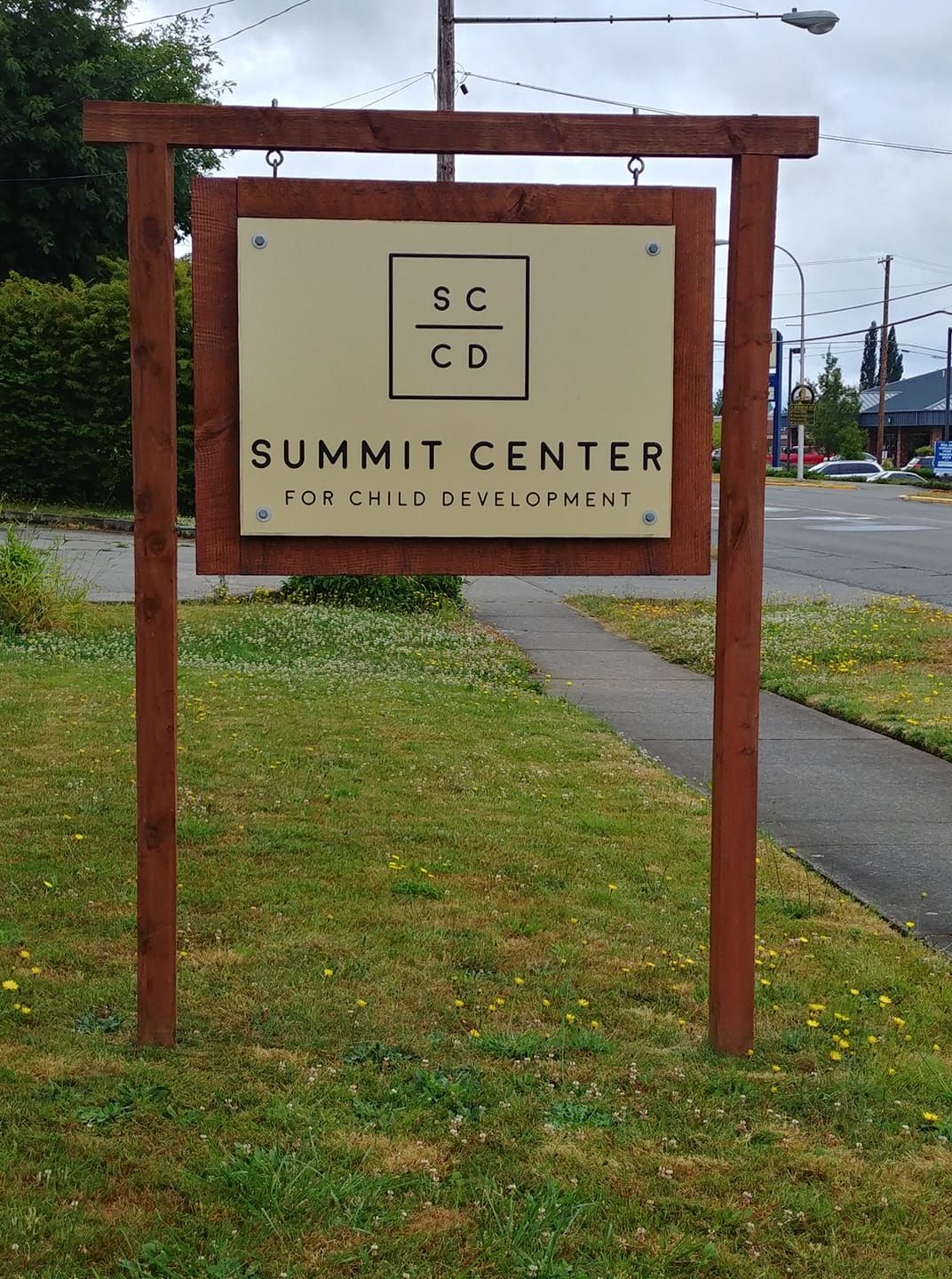 Wooden sign for Summit Center on a grassy roadside, with a sidewalk and trees in the background.