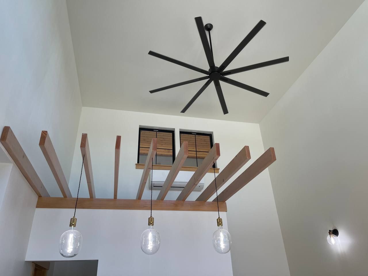 High ceiling with black ceiling fan and wooden hanging rack over pendant lights in a white room