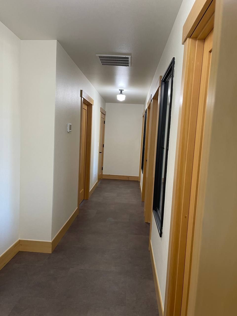 Narrow indoor hallway with white walls, wood doors, and a dark floor.