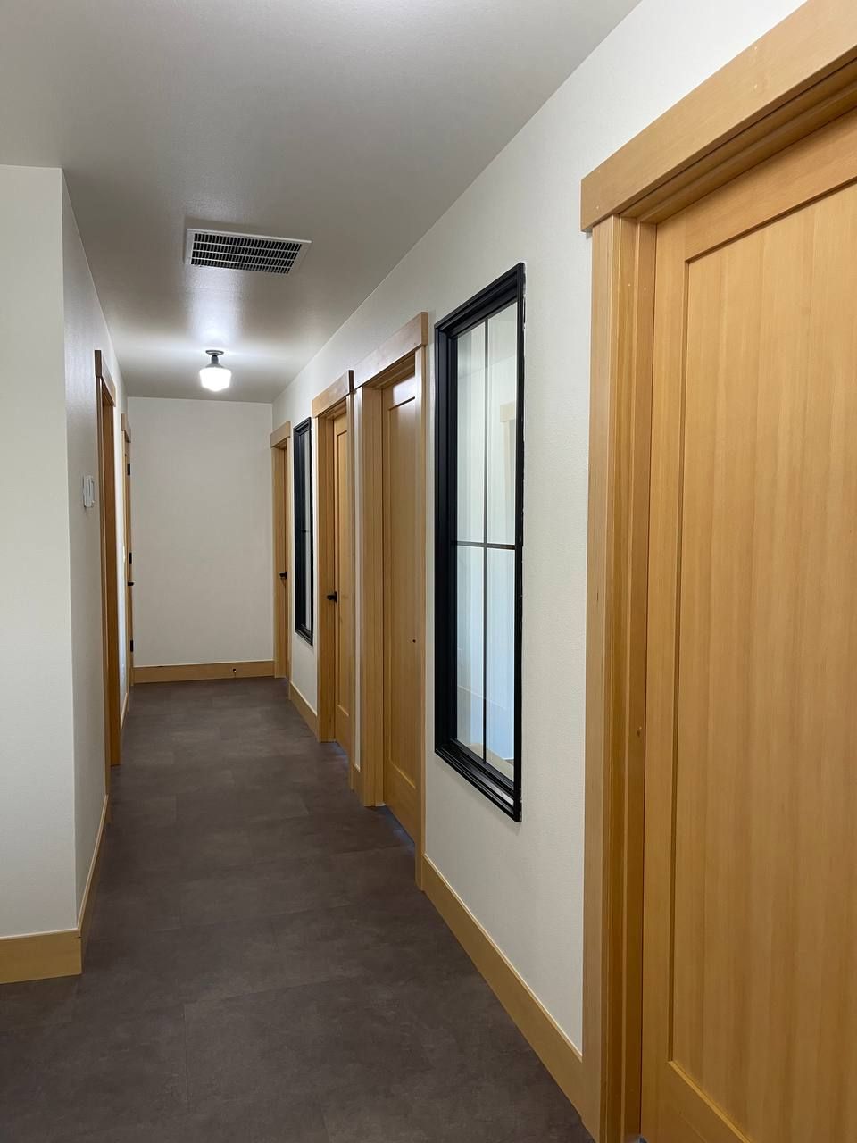 Narrow hallway with wood doors, white walls, dark floor, and frosted windows under ceiling lights