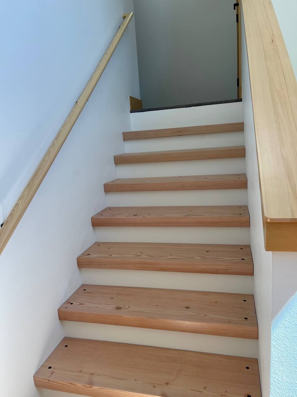Wooden staircase with handrails in a narrow indoor hallway, viewed from below.