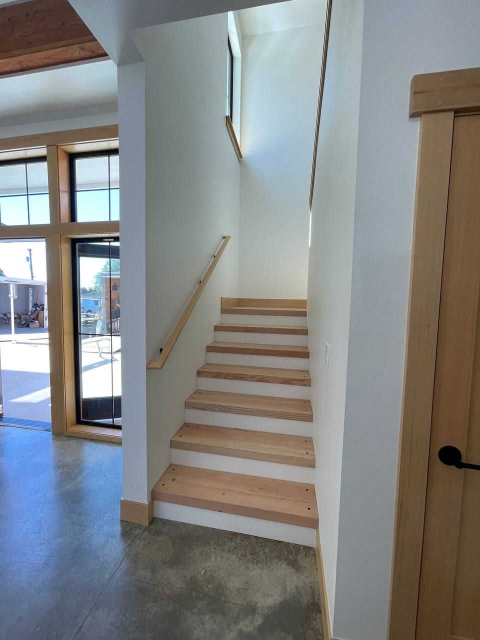 Indoor stairway with light wood steps, white walls, and a handrail beside an entryway.