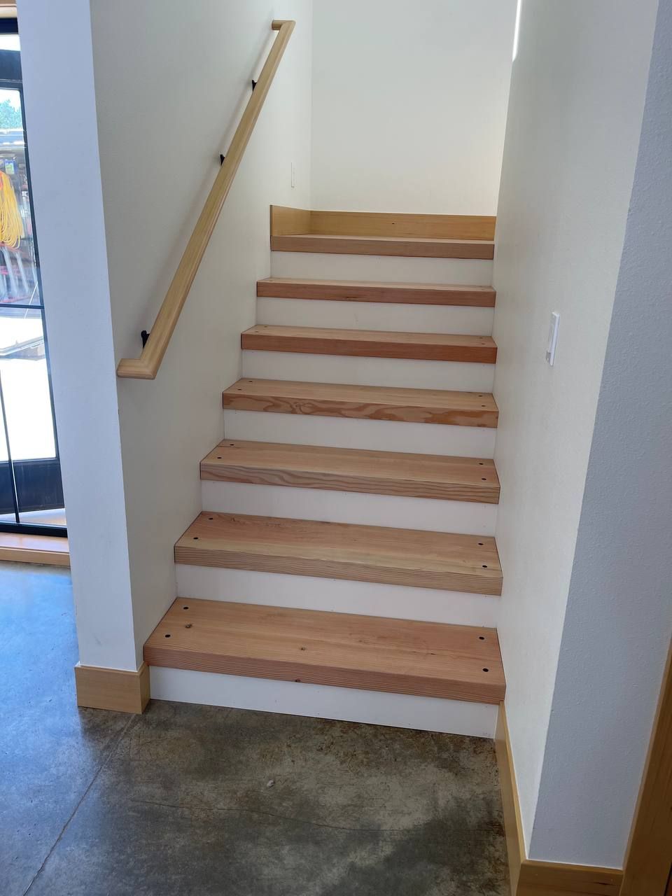 Indoor staircase with white walls, light wood handrail, and concrete floor landing.