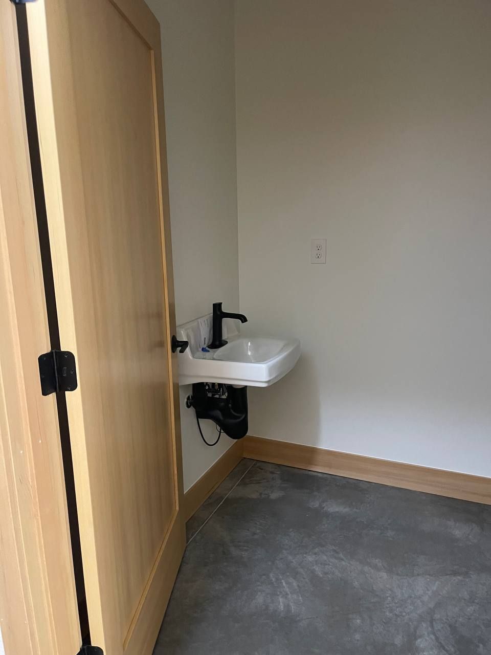 Small room with a sink and mirror on a wall, beside a wooden stall door and gray carpeted floor.