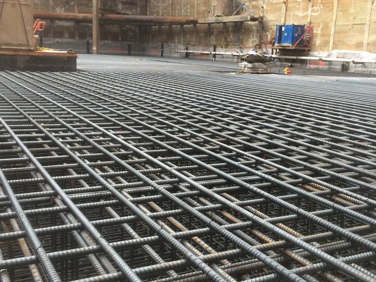Steel rebar grid laid across a construction foundation near a concrete wall and equipment