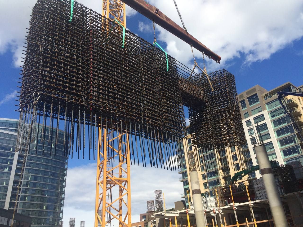 Construction site with a yellow crane lifting steel rebar panels amid high-rise buildings under a blue sky