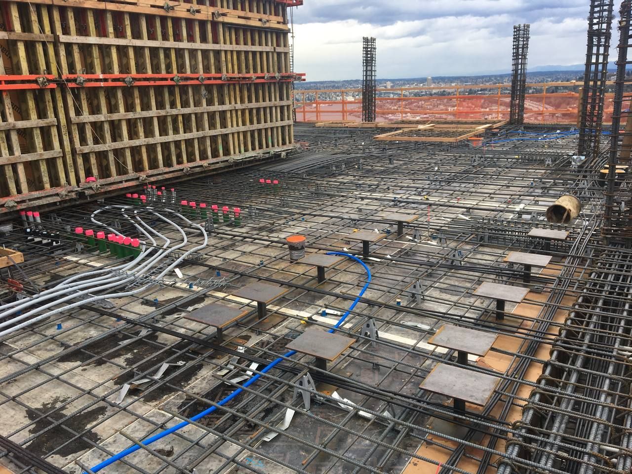 Construction site with dense rebar grid, wooden formwork, and blue conduit on a concrete slab under construction