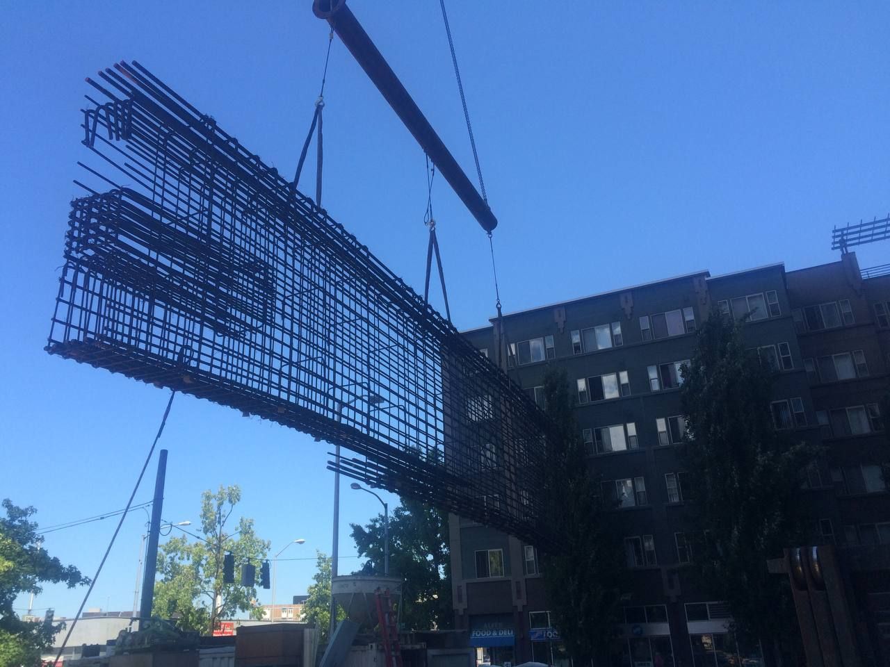 A crane hoists a large steel rebar frame beside a multi-story building under a clear blue sky.