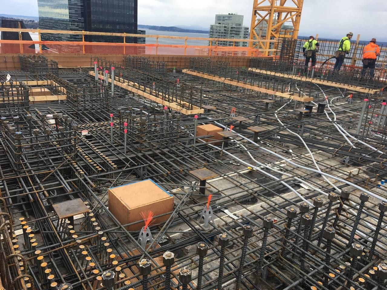 Construction site with rebar foundation, orange safety barriers, crane, and workers in the background