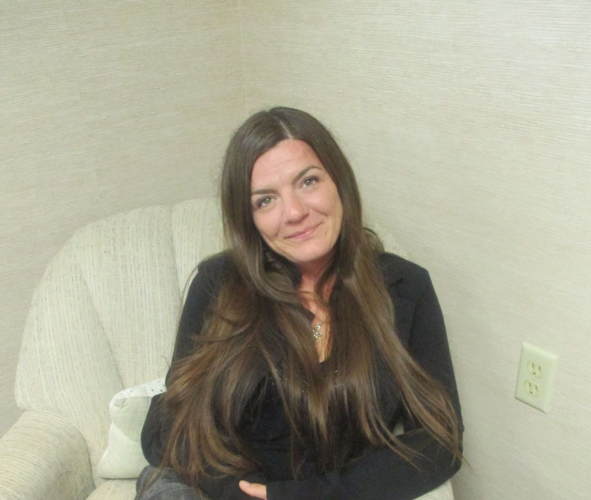 Woman with long brown hair smiles in an armchair against a tan wall.
