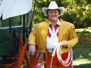 A man in cowboy attire, including a fringed jacket and hat, leans against a wagon, holding a rope.