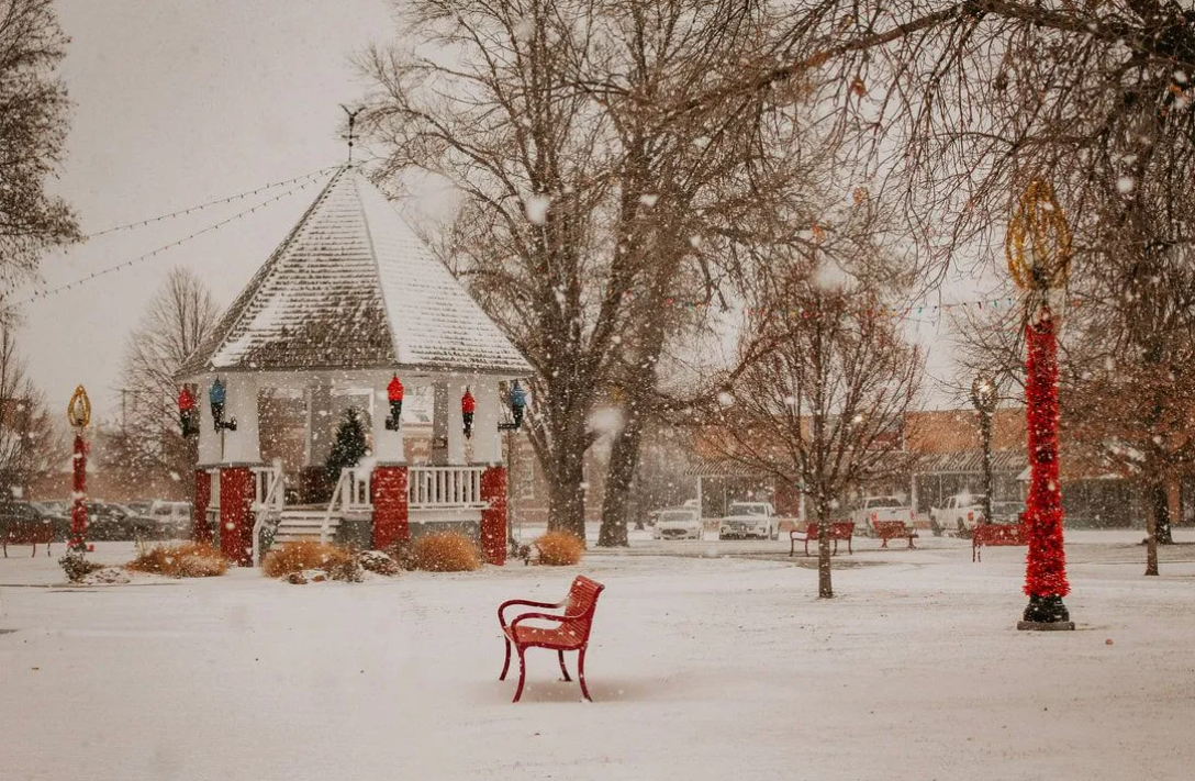 Snowy park with a gazebo, red accents, and falling snowflakes. A bench sits in the foreground.