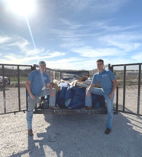 Two men in blue shirts posing with a large fish on a trailer in a sunny outdoor lot