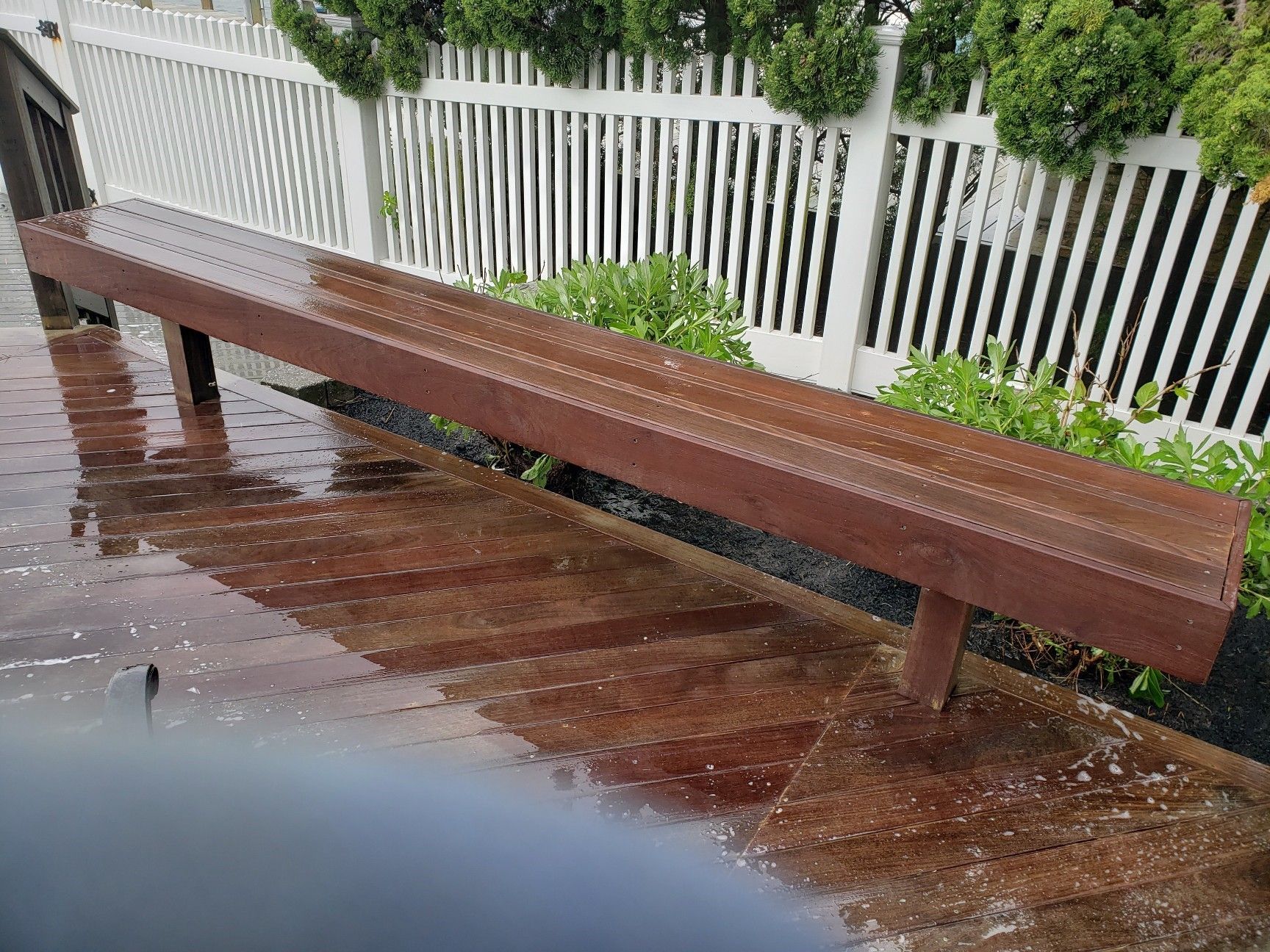 A wooden bench is sitting on a wet deck next to a white fence.