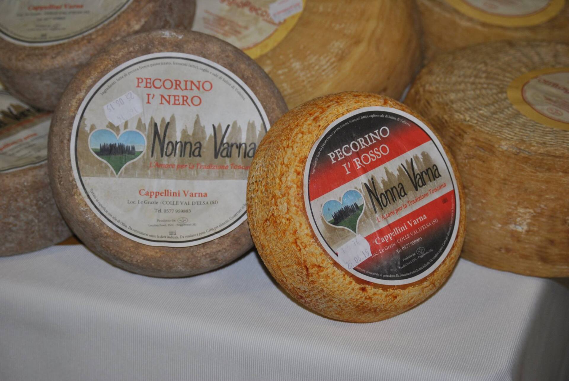 Forme di formaggio Pecorino con etichette, di varie dimensioni e colori, su una superficie bianca.