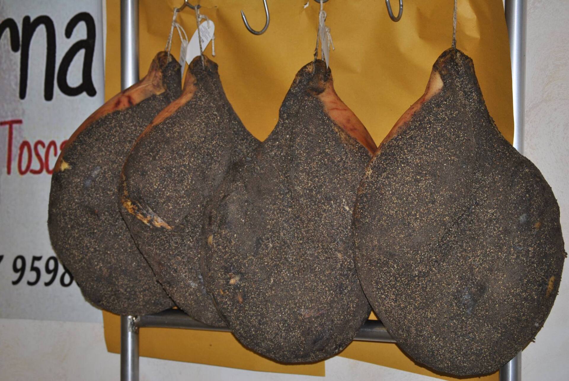 Quattro salumi stagionati, ricoperti di pepe nero, appesi a ganci di metallo.