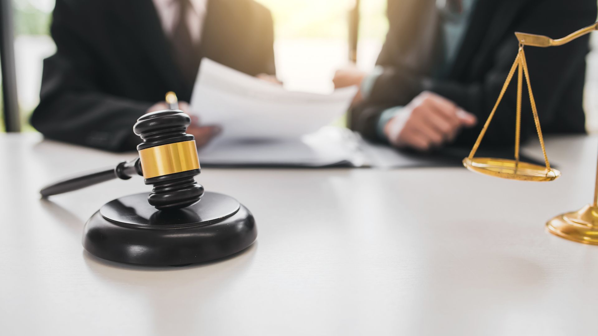 Gavel and balance scales on a table, with two people in suits reviewing documents in the background.