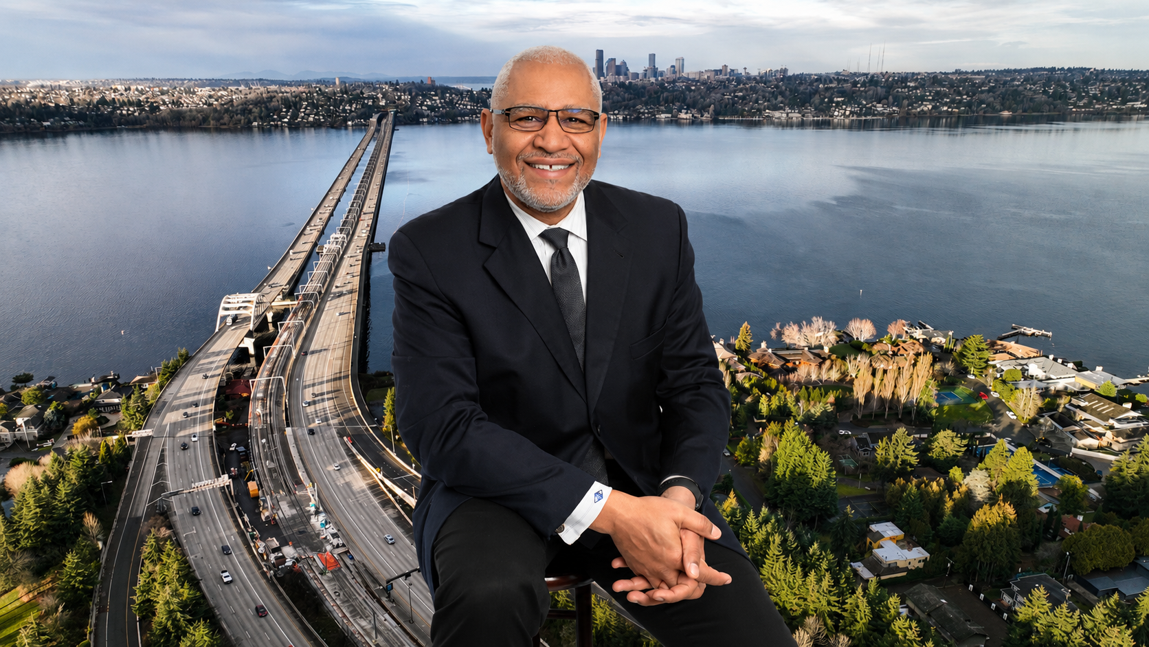 Smiling man in suit seated before a bridge and waterfront cityscape at sunset
