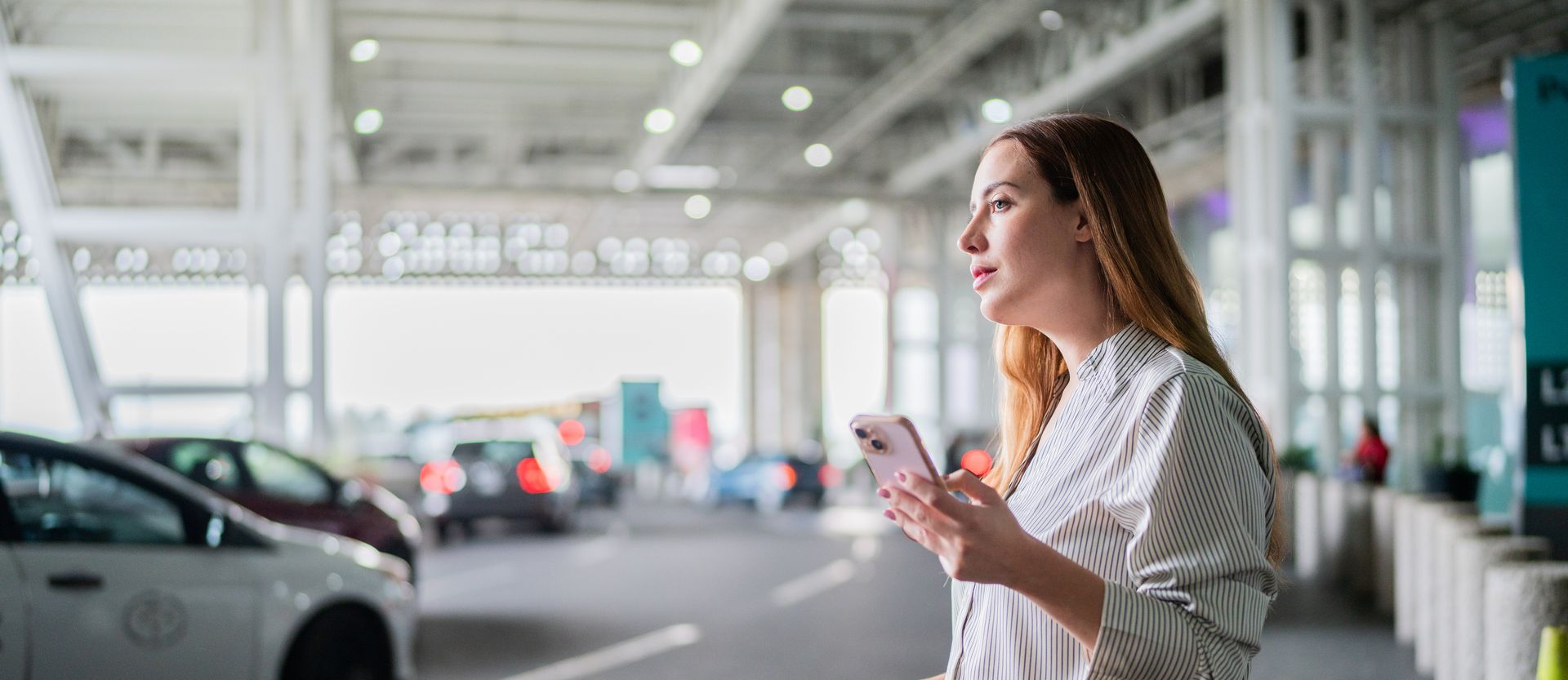 Woman holding a smartphone at an airport curbside terminal with cars and columns in the background