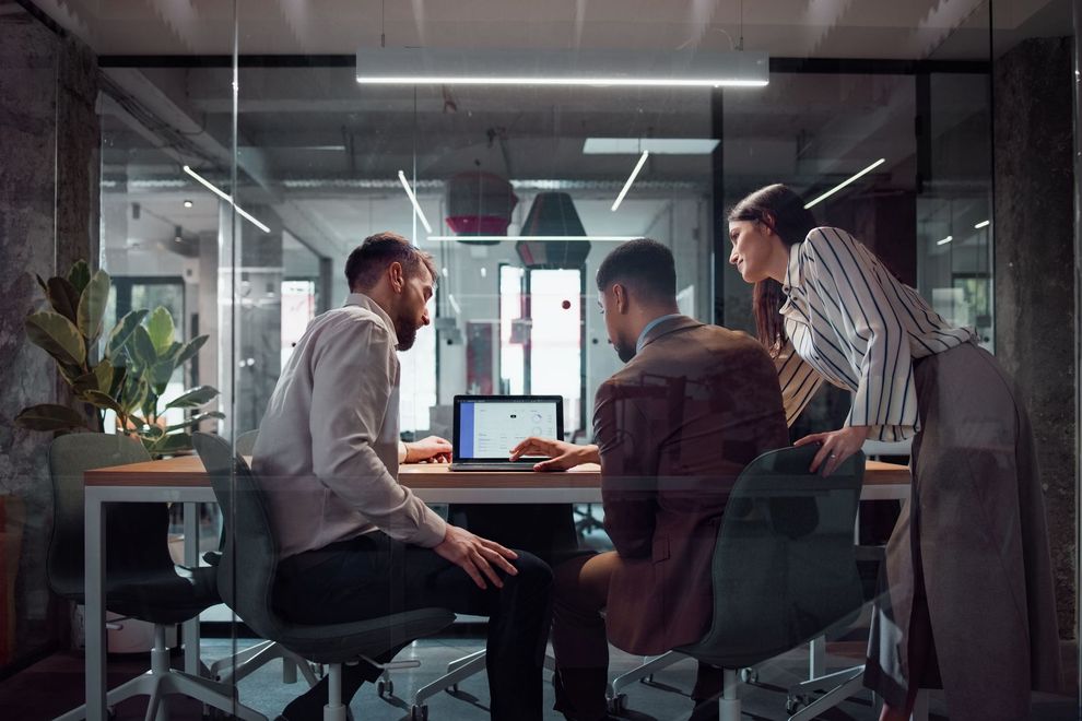 Three coworkers huddle around a laptop in a glass-walled office meeting room.