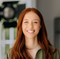 Smiling person with long red hair in a green shirt, standing indoors with a blurred background