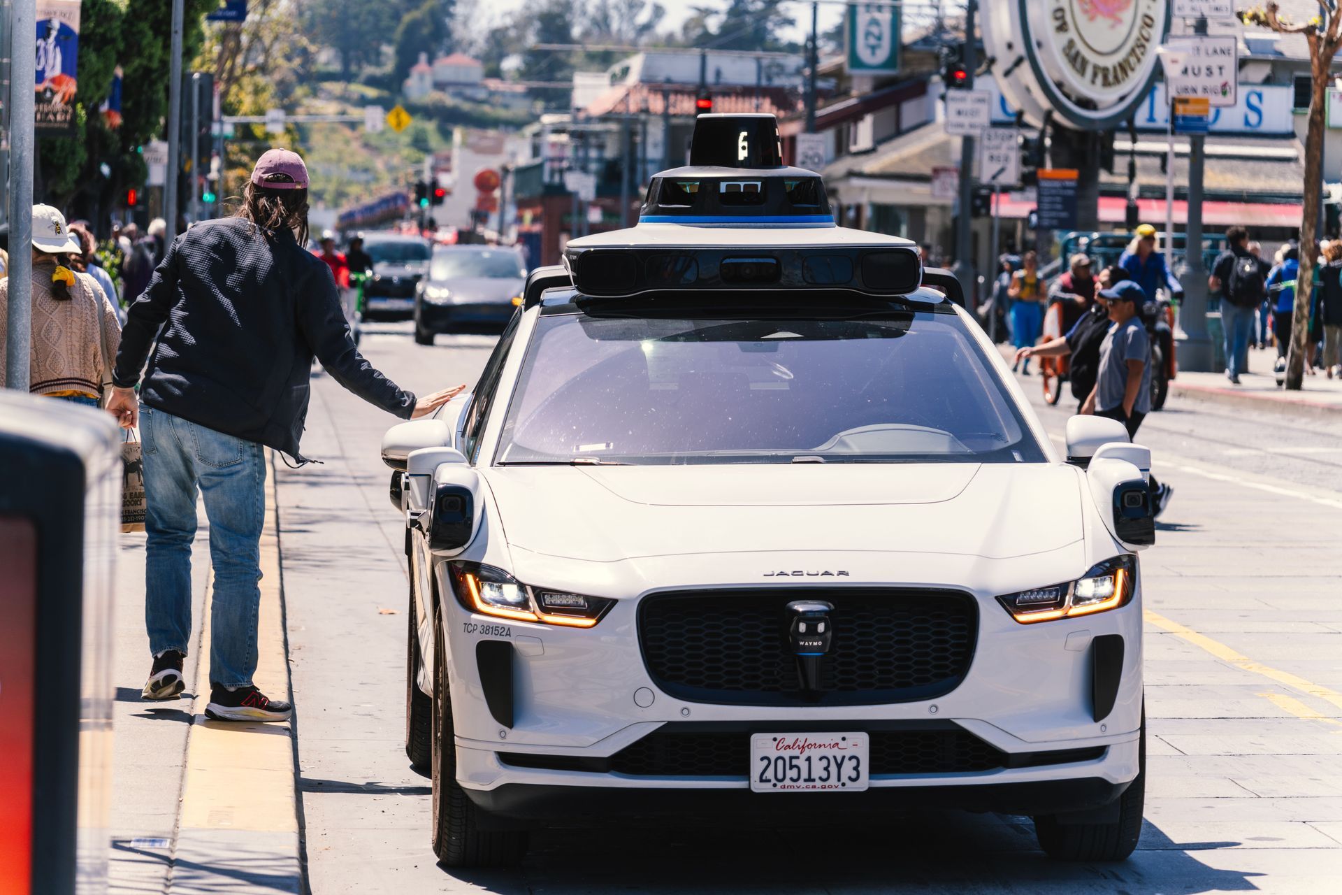 White self-driving car on a busy city street with pedestrians and storefronts in the background