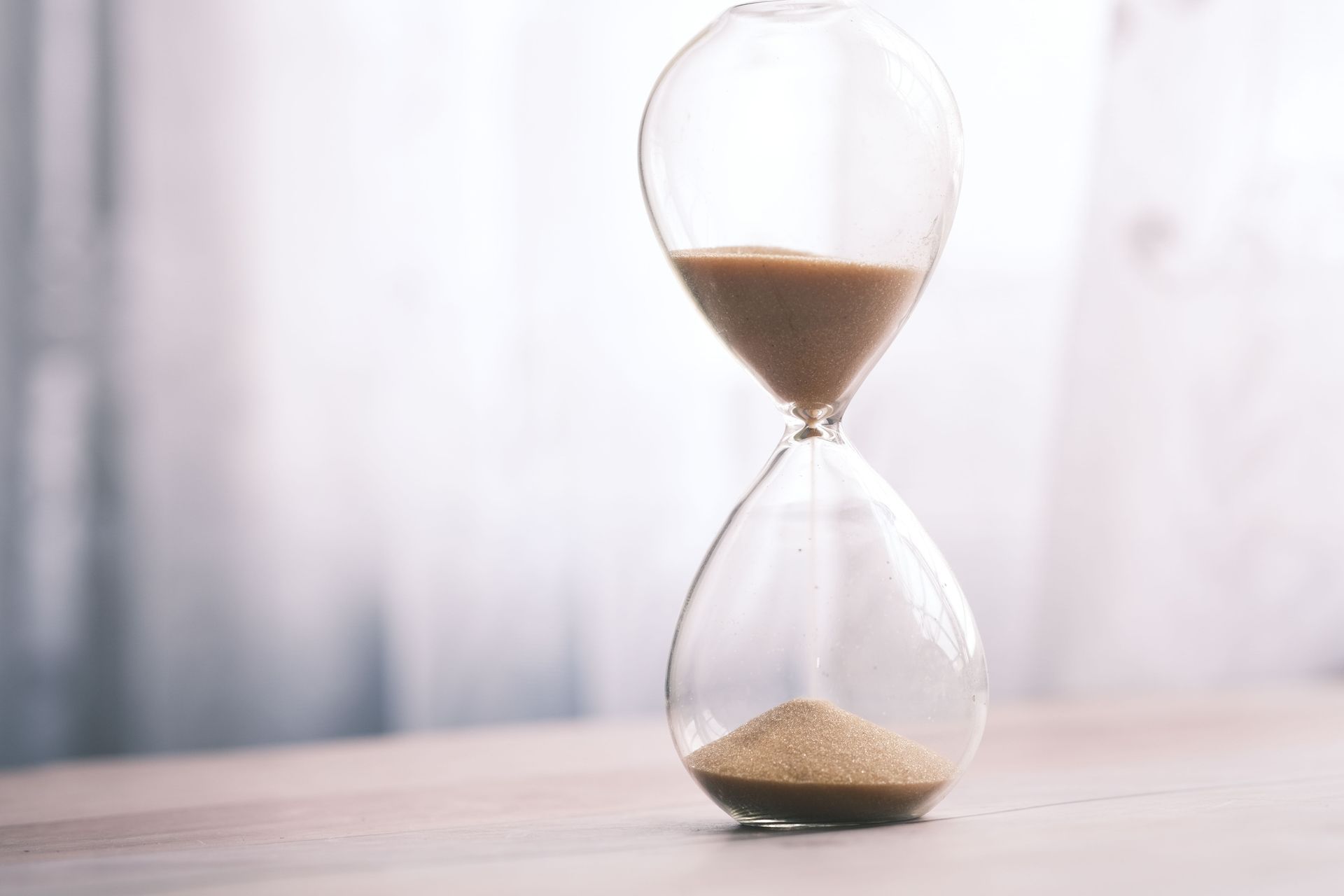 Hourglass with tan sand on a light wooden surface, half full, against a soft blurred background