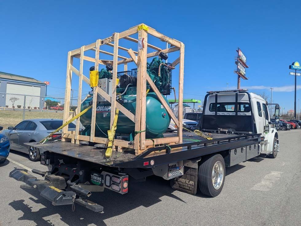 Large teal air compressor secured in a wooden crate on a flatbed tow truck. Sunny outdoor setting.