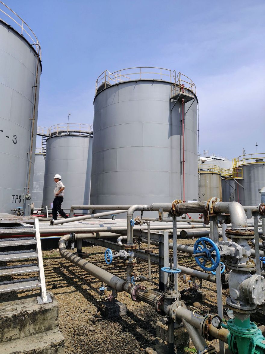 Industrial storage tanks and pipes at a refinery, with a worker in a hard hat walking near the tanks.