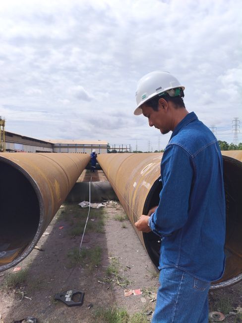 Worker in a blue shirt and hard hat inspecting large rusted pipes at an industrial site.
