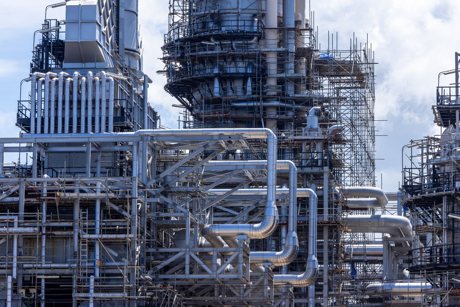 Large industrial refinery with metal piping, scaffolding, and tall processing towers under a cloudy sky